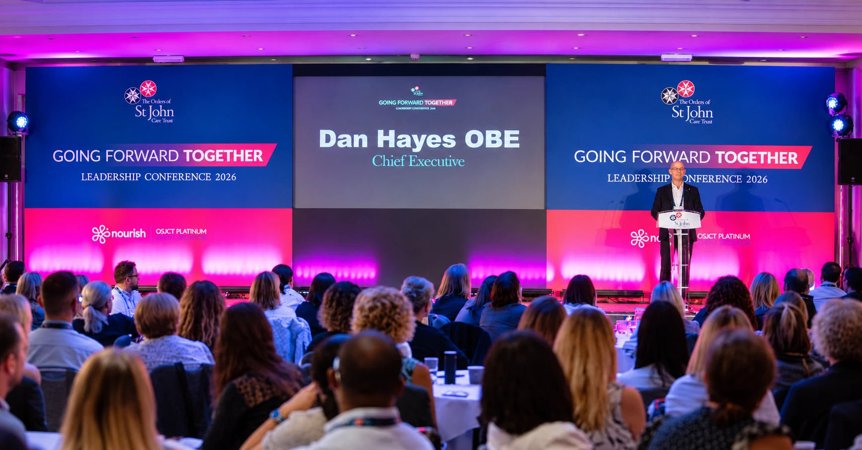 Dan Hayes OBE speaking at a lectern on stage at the St John Leadership Conference 2026, seen over a full audience